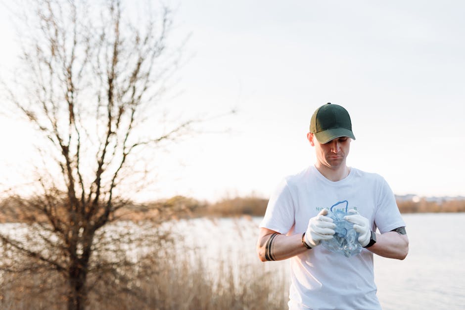 A young man wearing a white T-shirt, black baseball cap, and white gloves is standing outdoors near a body of water, holding a clear plastic bag filled with various litter items. The background featur