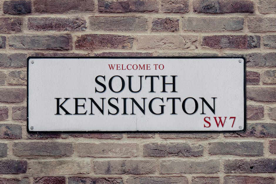 A rectangular metal street sign mounted on a brick wall displaying the text 'Welcome to South Kensington' with 'SW7' in red letters at the bottom right. The sign features a white background with black