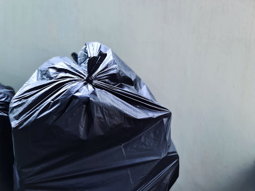Close-up of multiple black rubbish bags made from glossy plastic material, filled with waste and tightly tied at the top, placed against a plain, light-colored wall or backdrop. The bags are situated 